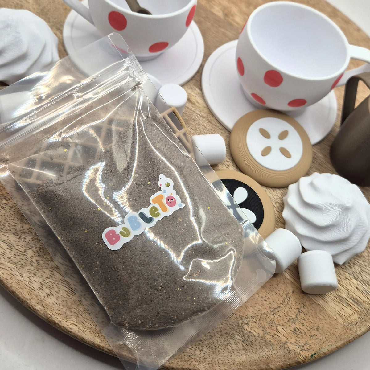 Priehľadné vrecko peniaceho prášku BubleTo s vôňou kakaa na podnose medzi šálkami s bodkami, koláčikmi a marshmallow